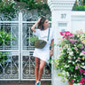 Woman in a white dress standing in front of a decorative white gate with floral arrangements wearing Elyse & i olive green crossbody bag. 
