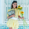 Woman in a yellow outfit holding sunflowers and a gray handbag against a decorative wall.