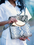 Person holding a patterned clutch bag with a blurred background putting a gold Bella purse into a bag. 